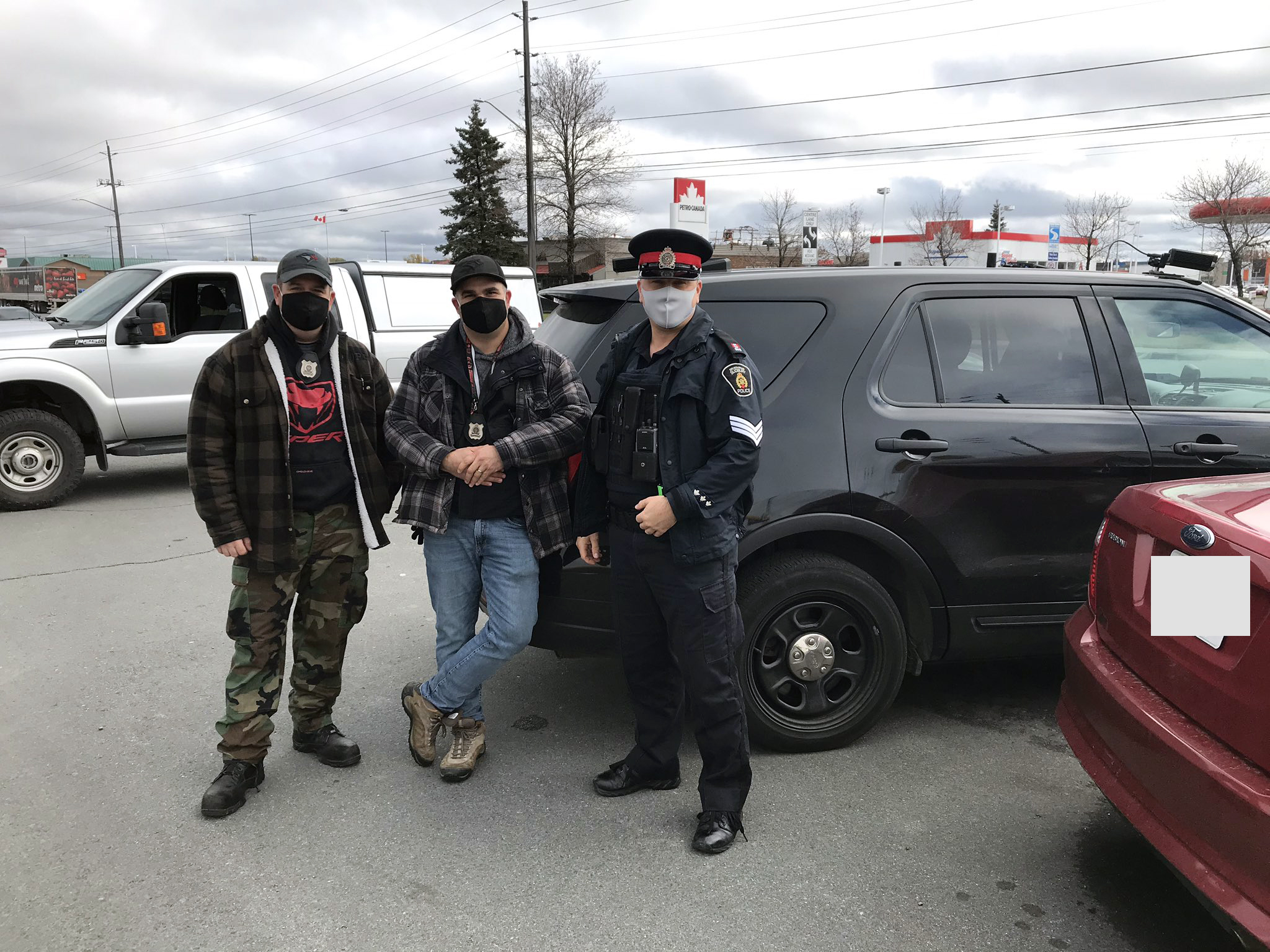 three officers in plain clothes posing in front of police cruisers outside